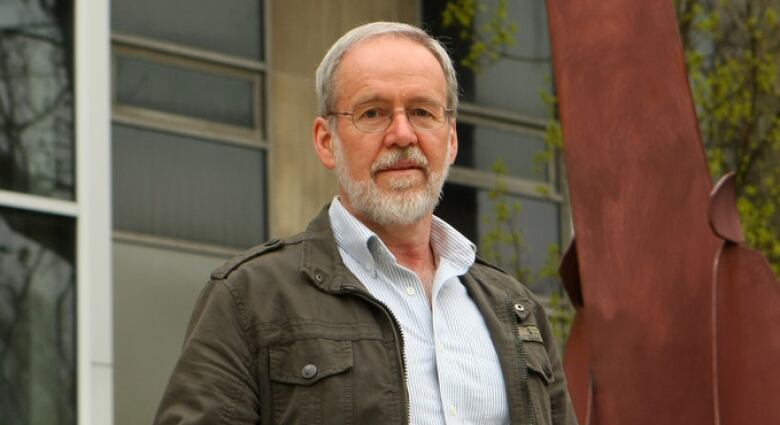 A man with grey hair and beard wearing glasses stands outside a building.