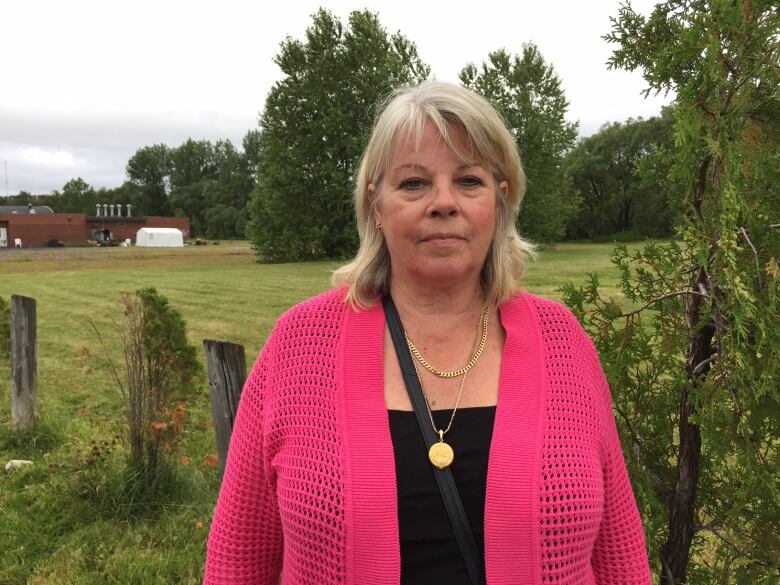 A woman with a pink sweater stands with grass and trees behind her.