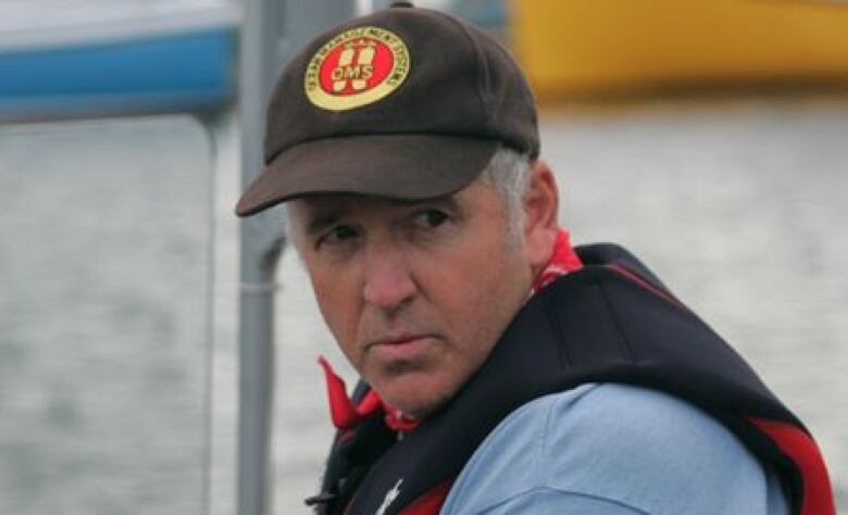 A man with a black hat looks over his left shoulder. He is wearing a black life jacket. There is water behind him.