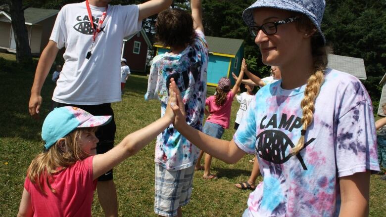 Kids give counsellors high fives outdoors at camp with cabins the background