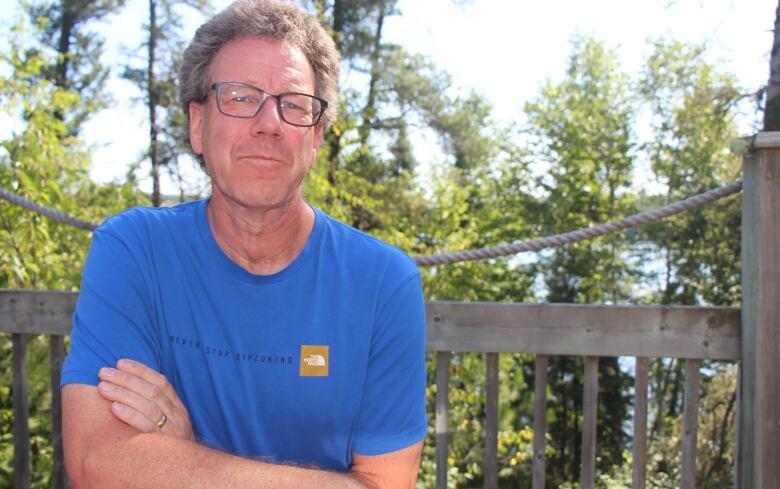 A man wearing glasses and a T-shirt sits outside with his arms crossed.