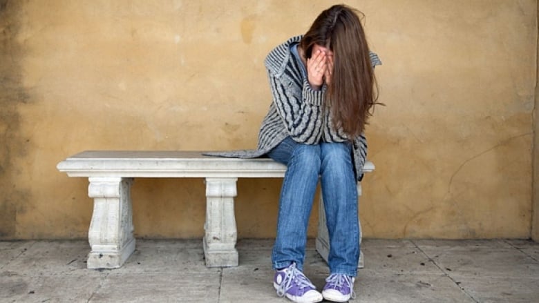 A young woman sits on a bench with her head in her hands.