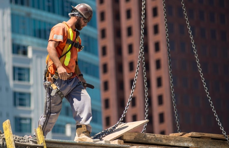 A man in a hard hat, reflective vest, work boots and safety harness is pictured in an elevated position on a building construction site.