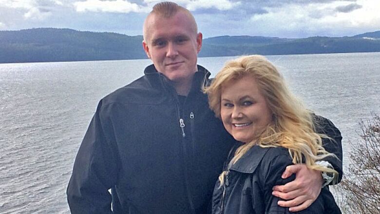 A young man in a black jacket has one arm over his mother, a woman in a black coat with long, blonde hair. They smile at a camera, behind them a body of water, mountains in the horizon.