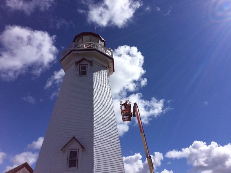 A person in a boom truck is painting a lighthouse. 