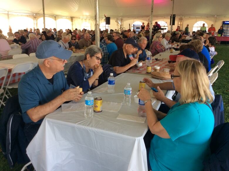 People eat lobster rolls as they sit at long tables in a tent. 