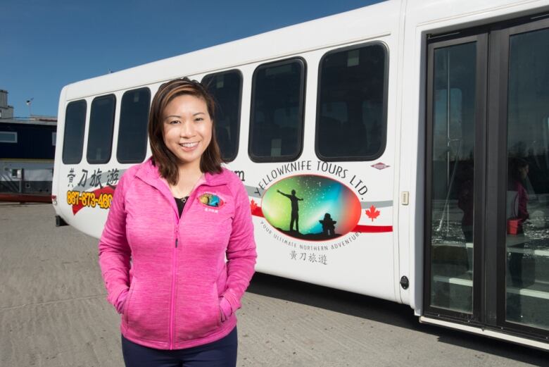 Woman in pink sweater poses in front of bus with Yellowknife Tours logo on it. 