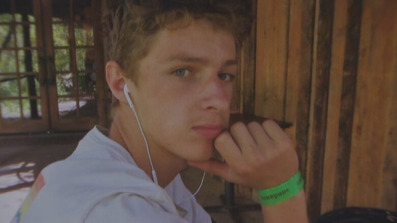 A young man with light brown curly hair wearing earbuds and a green wristband looks matter-of-factly into the camera.