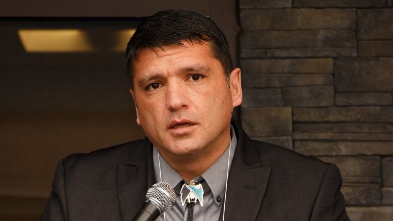 Man with dark brown hair and brown eyes wearing dark grey suit jacket and grey dress shirt sits in at table front of brick wall.