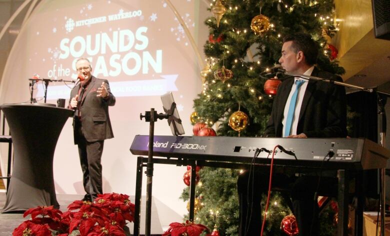 A man sits at a keyboard and speaks to the male host of the show who is on the stage a short distance away