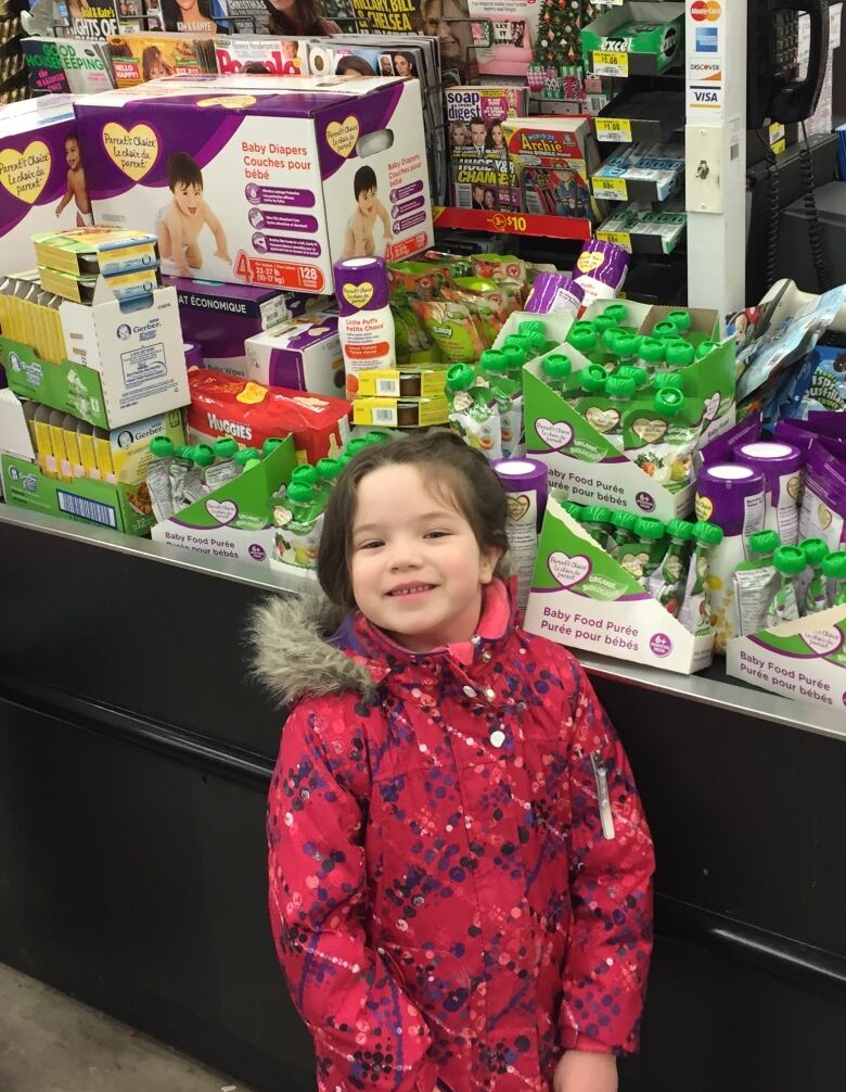 A young girl stands in front of a collection of baby food, wipes and formula.