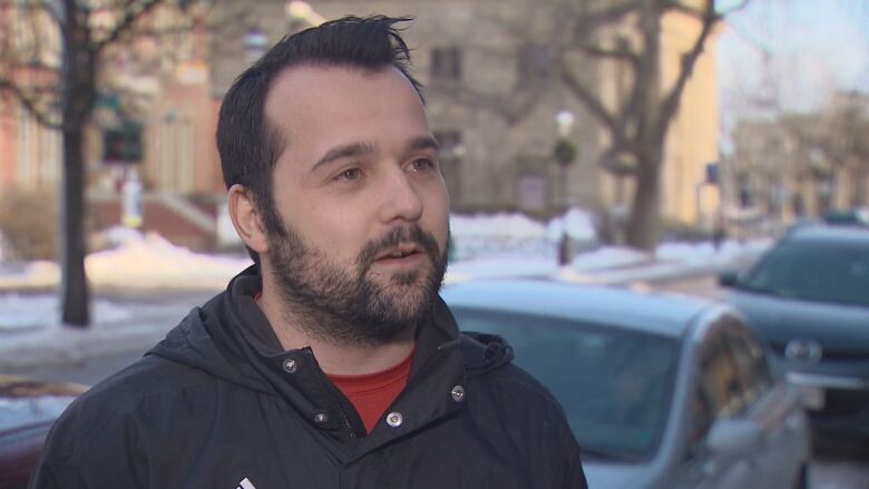 A man with a beard stands outside wearing a jacket with snow on the ground in the background.