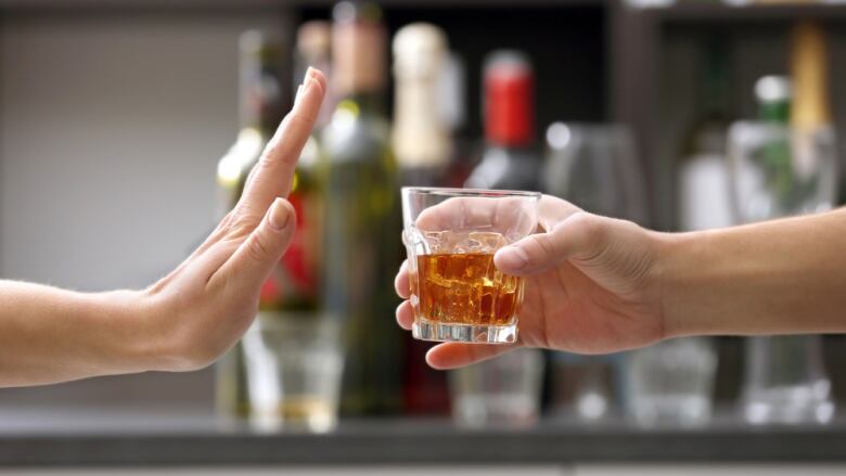 A person holds up her hand as if to stay stop while another hand offers a drink with a range of alcohol blurred out on a shelf behind as if in a bar.