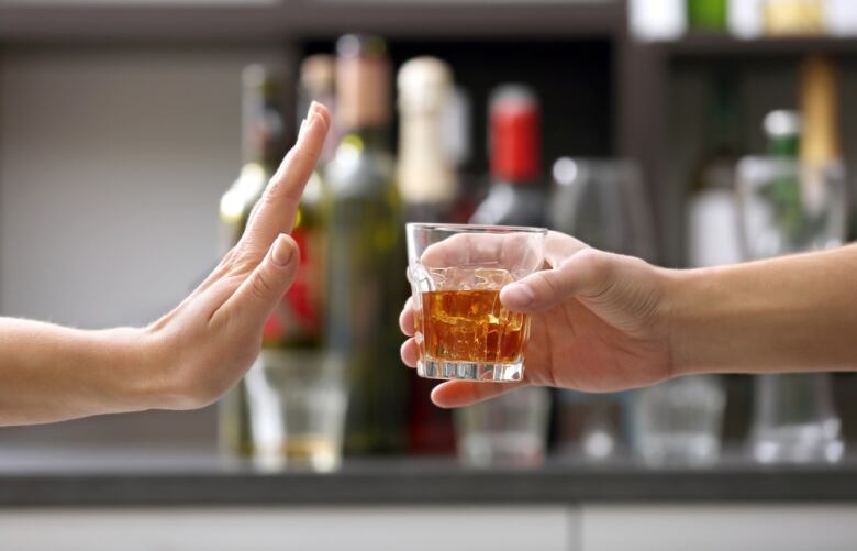 A person holds up her hand as if to stay stop while another hand offers a drink with a range of alcohol blurred out on a shelf behind as if in a bar.