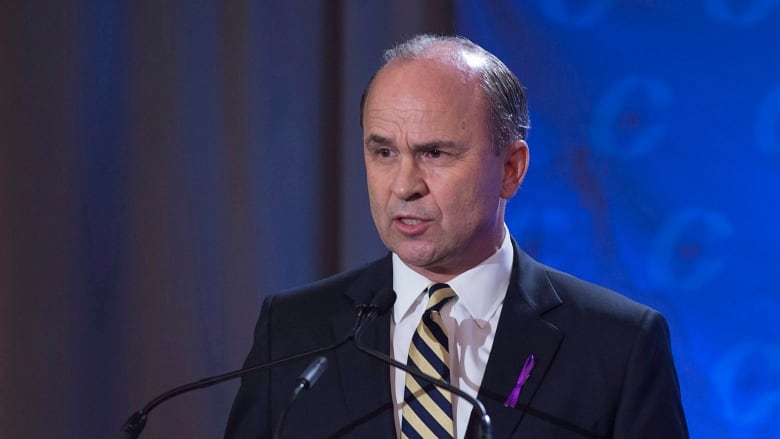 A man in a dark suit stands at a podium in front of a blue background.