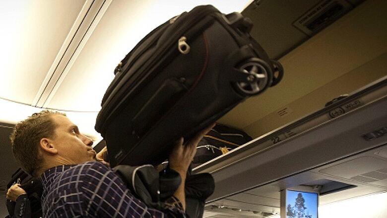 A man putting a carry-on suitcase in the overhead compartment on a plane. 