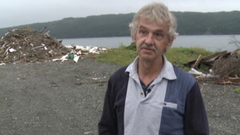 A thin grey-haired man is in the foreground. He is standing in front of a body of water. 