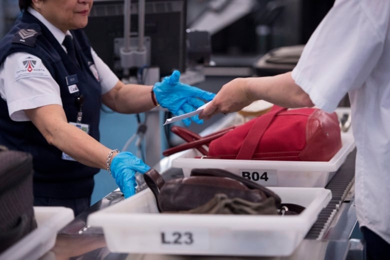 Security officer at an airport wears gloves and monitors travellers as they put their luggage in big plastic bins, which move down a conveyor belt.