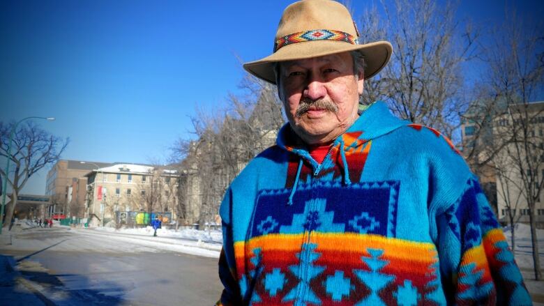 A man with a moustache stands outside in a colourful coat, wearing a hat 