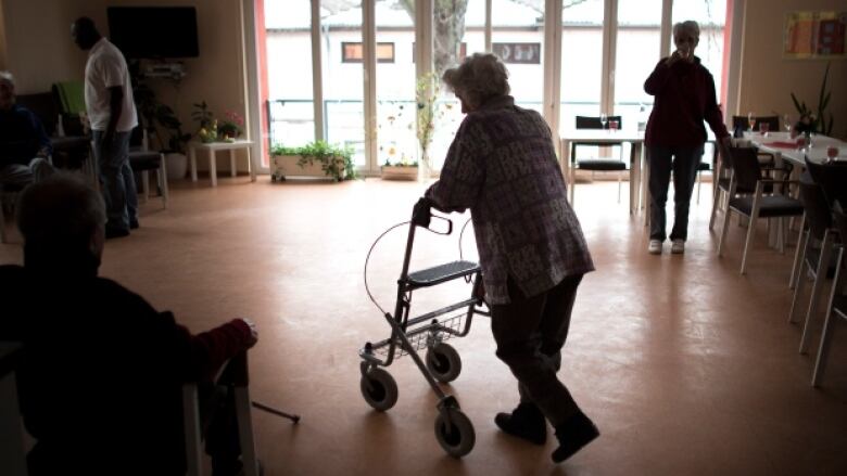 An older woman uses a walker in a room with a large window, dining tables to the right and chairs to the left.