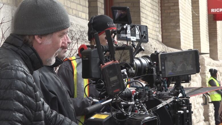 People wearing touques and winter coats stand behind cameras on a city street.