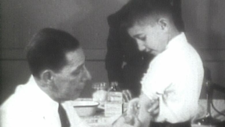 An old black and white image of a doctor, kneeling down, and injecting a needle with the polio vaccine into the arm of a young boy, standing in front of him.