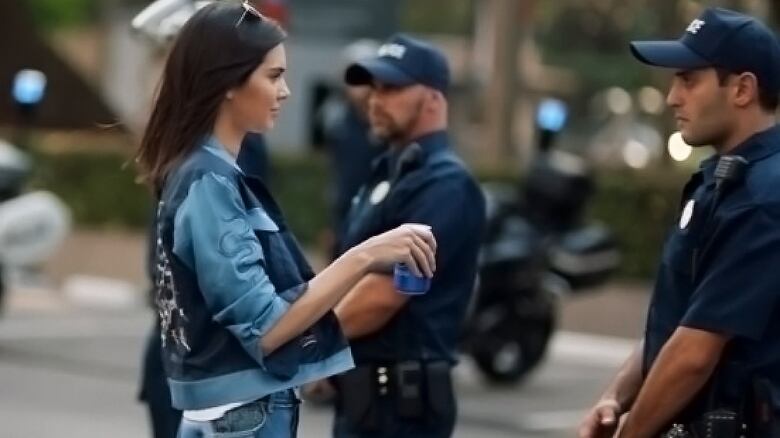 A woman offers a can of Pepsi to one of several police officers standing in a line.