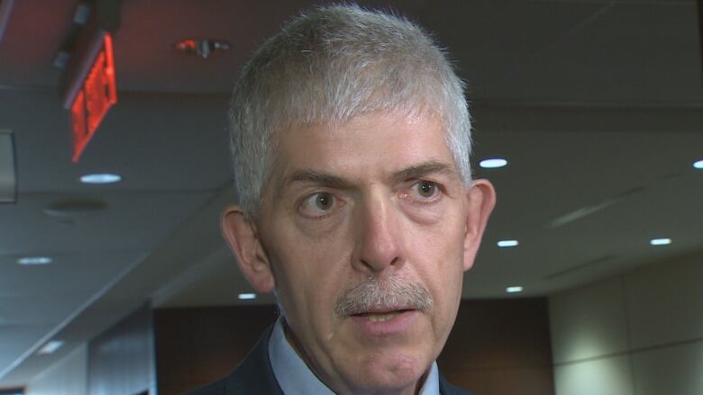 A man with short gray hair and a thin mustache looks slightly off to the right while facing the camera in a head and shoulders image. He's wearing a light blue dress shirt, and a dark blue suit. The background is an office building corridor or lobby.