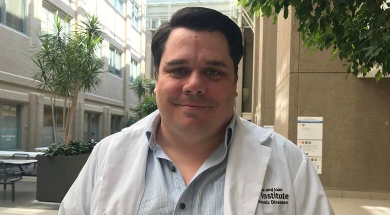 Craig Jenne wears a white lab coat and is standing in an atrium, looking directly into the camera.