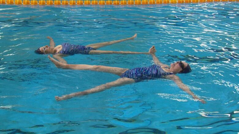 Two women, wearing blue one-piece swimming suits, goggles and swim caps, are floating on their backs in a swimming pool.