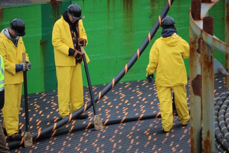 Three workers dressed in yellow rain gear with black hard hats stand on a ship handling cable being installed on the sea floor. 