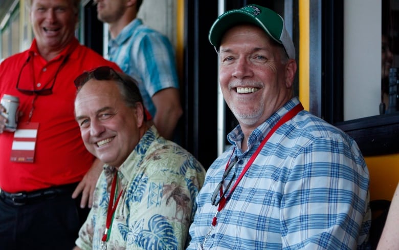 Two men in casual attire sit next to each other in the bleachers of a sports game, smiling at the camera.