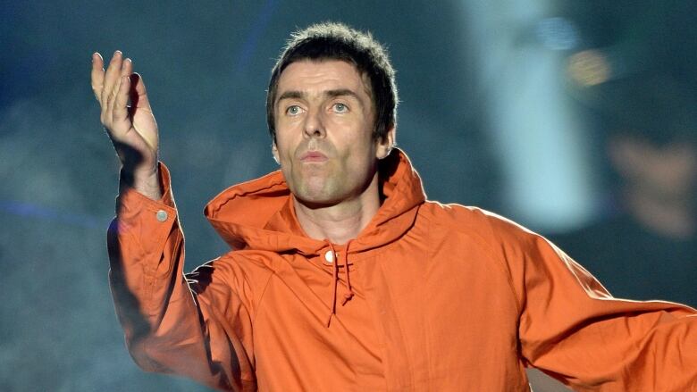 Liam Gallagher onstage in a long orange raincoat and black pants