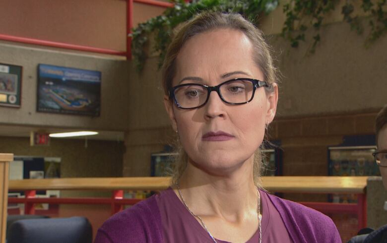 A woman wearing glasses and a purple sweater looks away from the camera while standing inside a building with brown walls behind her.