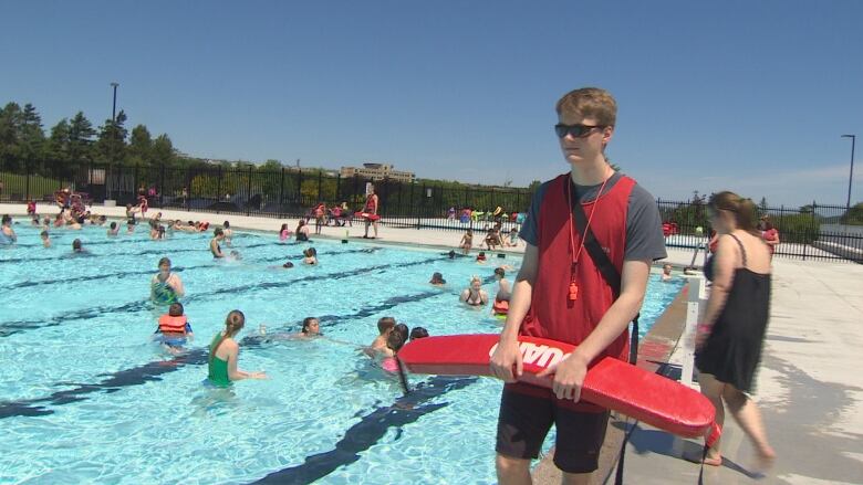 A life guard wearing red stands next to a swimming pool. 