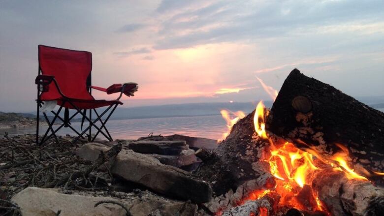 An empty red camping chair sits in front of a burning campfire.