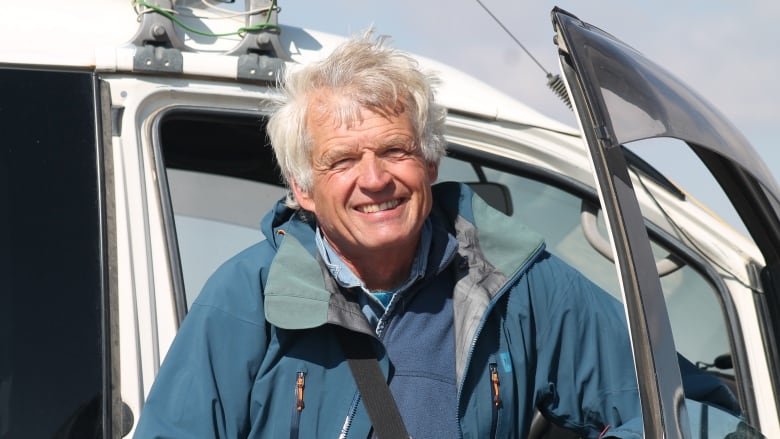 A white man with short white hair is wearing a blue jacket. He is smilling, while leaning in the doorway of a helicopter.