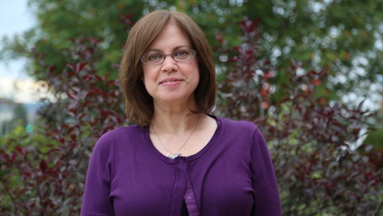 Woman with above shoulder length hair, wearing purple shirt, smiling at camera with a bush in the background.