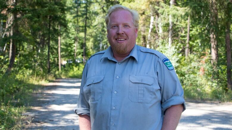 A man with a light red beard and blond hair in a blue open-collared shirt smiles at the camera framed by the forest behind him.