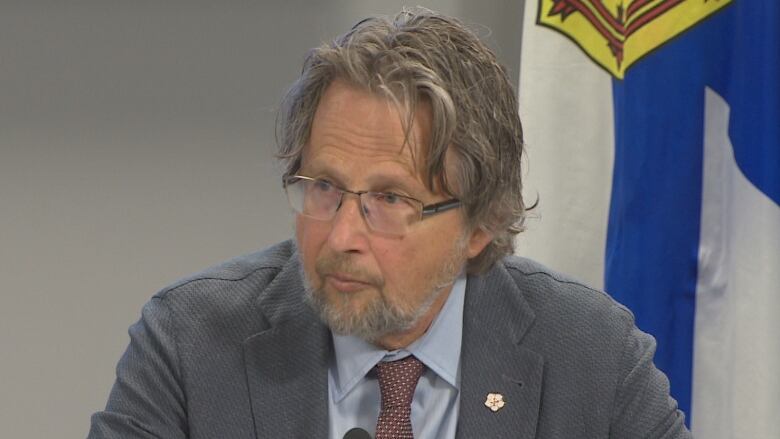 A man wearing glasses looks left and leans toward a small microphone. A Nova Scotia flag can be seen behind him.