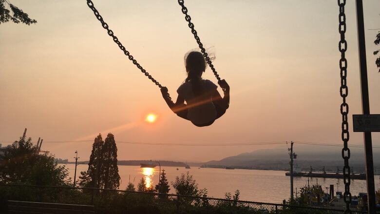 A silhouette of a young girl on a swing against a backdrop of Burrard Inlet with orange skies due to wildfire smoke and sunset.