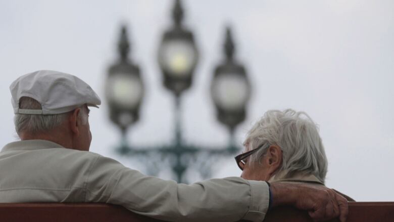 Old man and a woman with their back turned to the camera sitting on a bench overlooking a blurred street lamp.