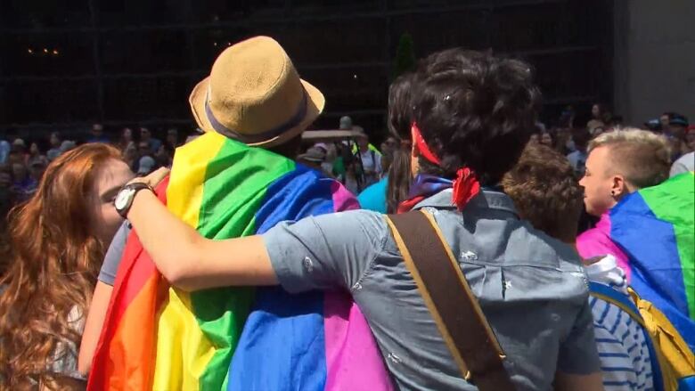 A crowd of people are seen from the back, some with multi-coloured Pride flags draped on their backs