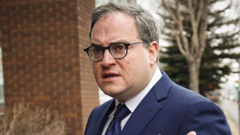 Man with glasses in blue suit gestures outside brown brick building.