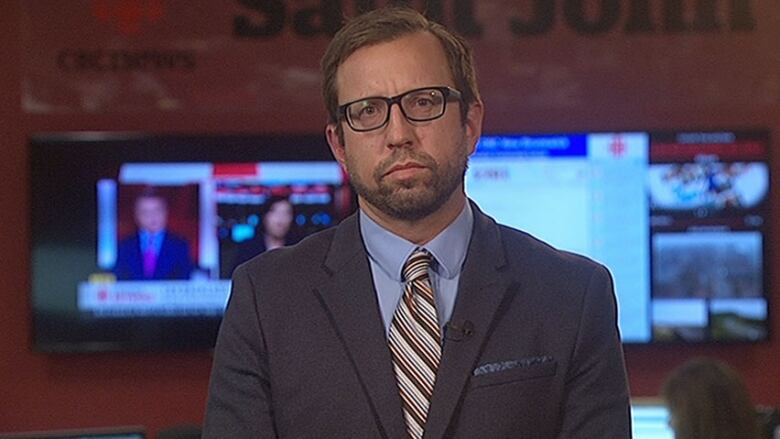 A man in a suit and tie sits facing a camera in a TV studio.