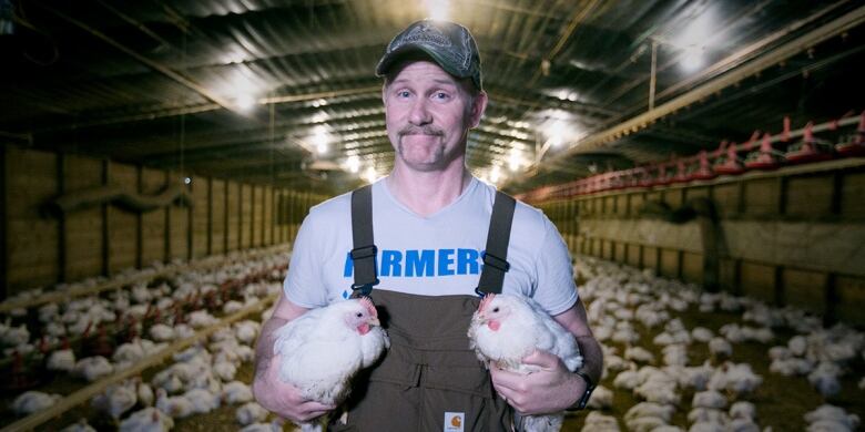 A man in overalls holds two chickens.