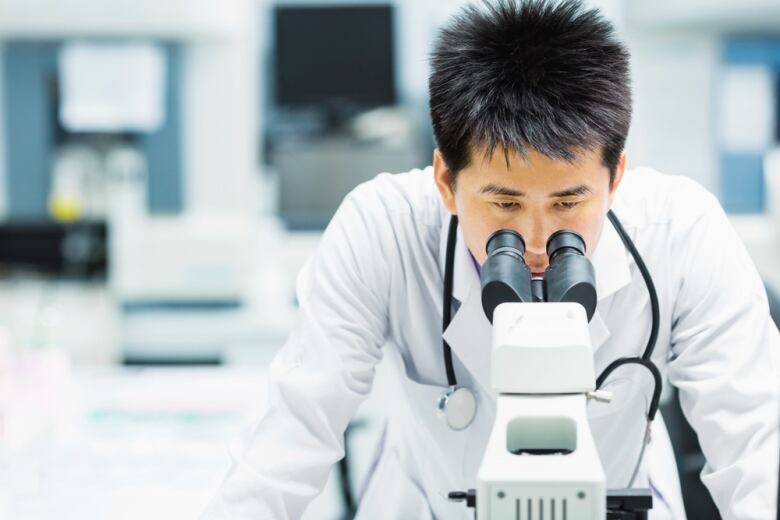 A  man wearing a white lab coat and stethoscope around his neck peers into a microscope.