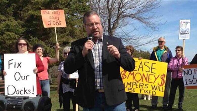 A man holding a microphone with protestors with signs in the background.