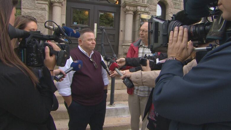 A person stands in front of a building, surrounded by people with cameras and microphones.