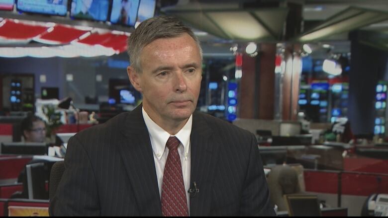 A white man with greying brown hair is seen wearing a black pinstripe suit and a red patterned tie.